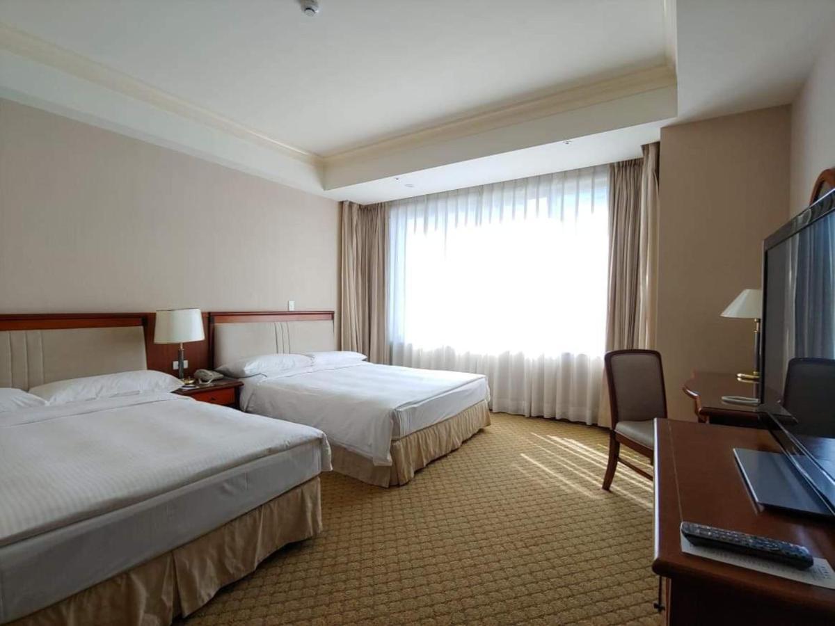 Evergreen Laurel Hotel - Taichung Exterior foto The image shows a hotel room featuring two single beds with white bedding. There is a large window with sheer curtains letting in natural light, and the walls are painted in neutral tones. A wooden desk with a chair is positioned near the window, and