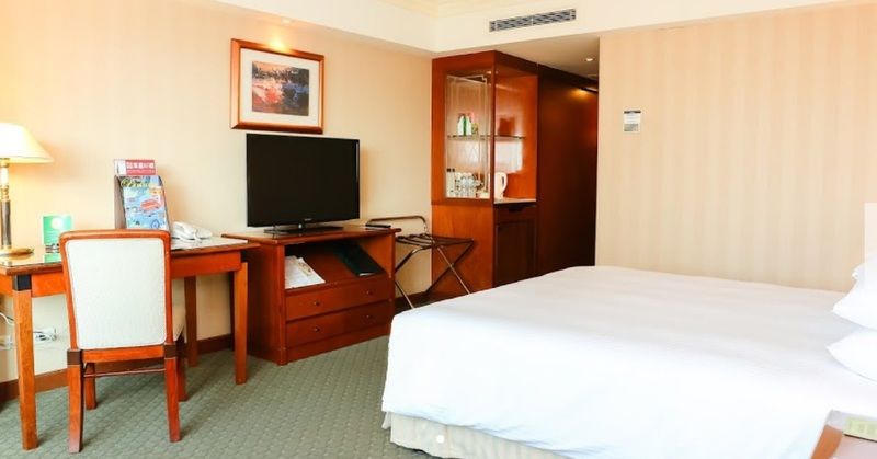 Evergreen Laurel Hotel - Taichung Exterior foto The photo shows a hotel room featuring a neatly made bed with a white blanket. On one side, there is a wooden desk with a chair, and a flat-screen television is mounted nearby. An accent cabinet is visible, displaying various items. The walls are pai
