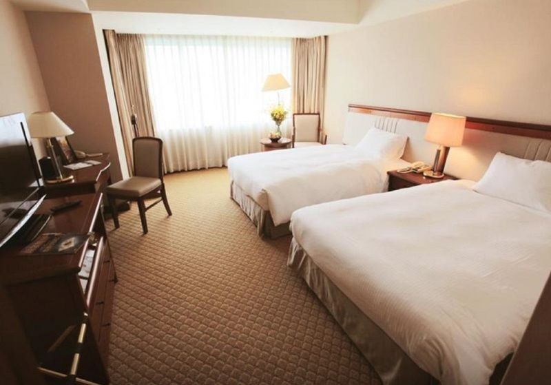 Evergreen Laurel Hotel - Taichung Exterior foto The photo shows a hotel room featuring two twin beds with white bedding. There's a chair positioned near one of the beds and a desk with a computer setup against one wall. Large windows allow natural light to enter the room, and light-colored curtain