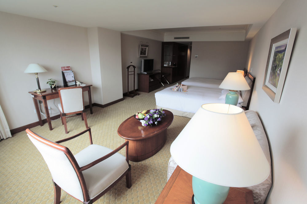 Evergreen Laurel Hotel - Taichung Exterior foto The photo shows an interior view of a hotel room. It features two beds, a coffee table with a small decorative arrangement of flowers, and a comfortable chair. There's a desk and a lamp, along with another larger lamp on the table. In the background,