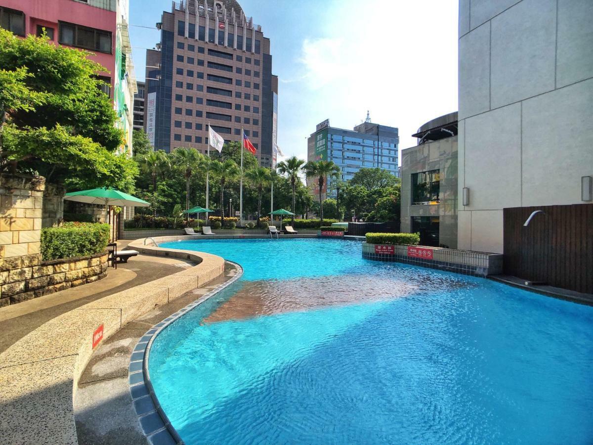 Evergreen Laurel Hotel - Taichung Exterior foto The photo shows a swimming pool set in a modern urban environment. The pool has a curved edge and is surrounded by greenery, including trees and umbrellas for shade. In the background, there are several tall buildings, indicating a cityscape, with so