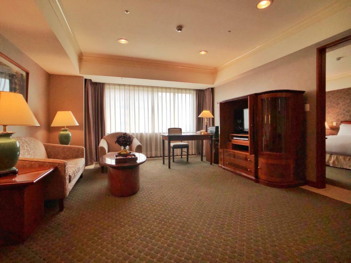 Evergreen Laurel Hotel - Taichung Exterior foto The photo shows a cozy hotel suite or living area. There is a sofa and an armchair arranged around a small coffee table. A desk with a chair is positioned near a large window covered with sheer curtains that allow natural light to enter the room. Two
