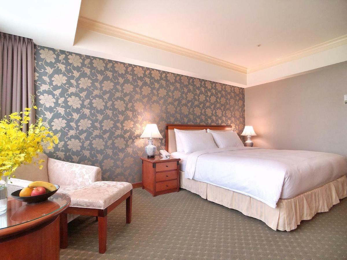 Evergreen Laurel Hotel - Taichung Exterior foto The photo shows a well-decorated hotel room. It features a large bed with white bedding, situated against a patterned wall with floral designs. On either side of the bed are bedside tables with lamps. There is a small seating area that includes a cha