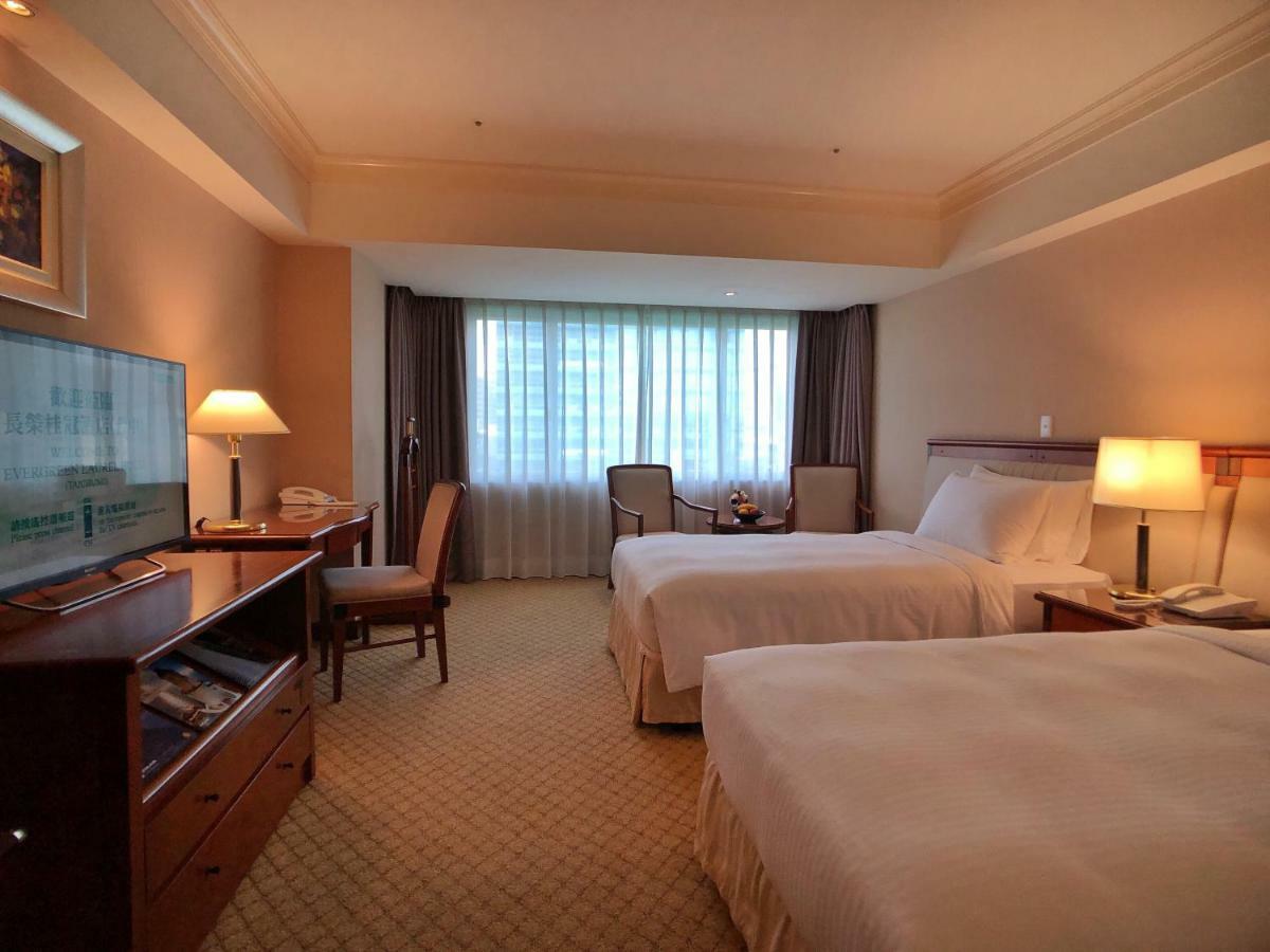 Evergreen Laurel Hotel - Taichung Exterior foto The photo shows a hotel room featuring two twin beds with white bedding. There's a television mounted on a wooden dresser, and next to it, a comfortable chair and a small table are positioned. The room has large windows that allow natural light to en
