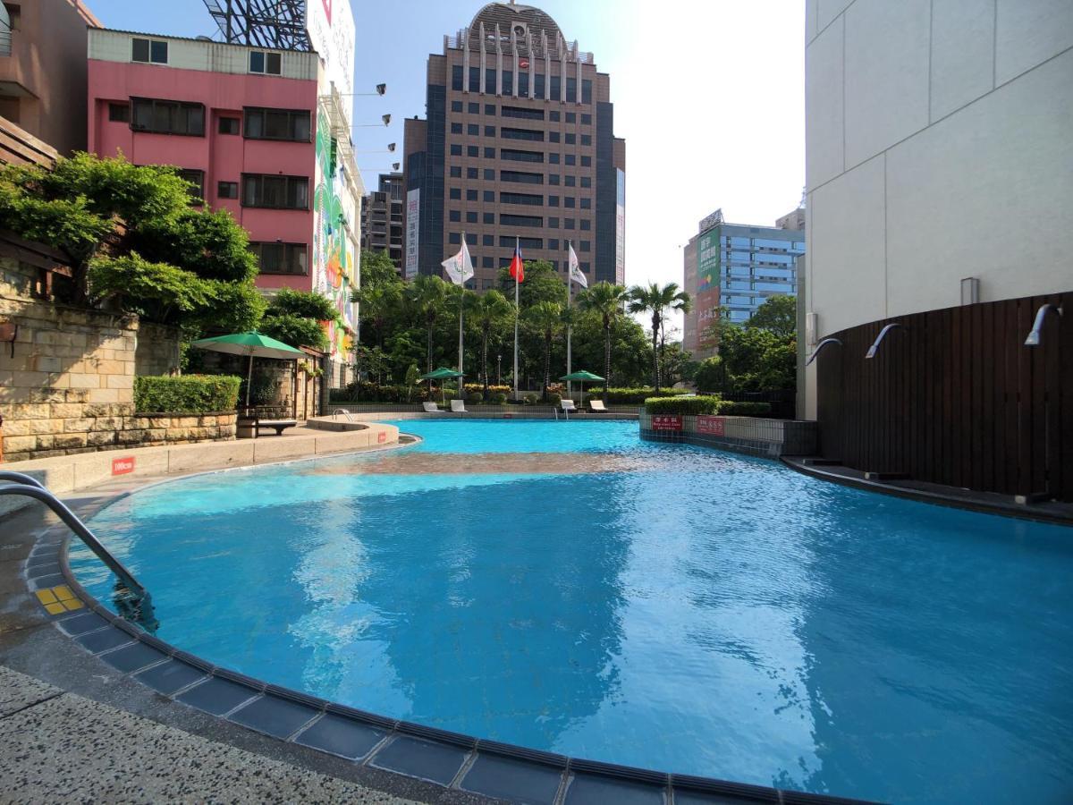 Evergreen Laurel Hotel - Taichung Exterior foto The photo shows a swimming pool area set in an outdoor environment. The pool has a clear blue water and a gently curving shape. Surrounding the pool are some green trees and plants, along with a few umbrellas providing shade. In the background, there